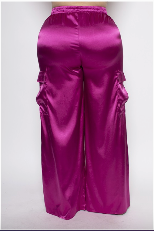 The back of a woman wearing a pair of vibrant pink, high-waisted satin plus sized cargo pants with functional pockets and an adjustable drawstring waist.