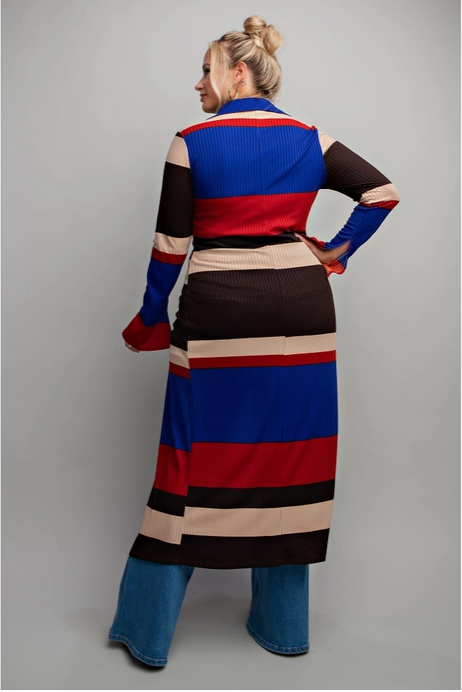 Woman wearing a longline, colorblock maxi shirt with bold horizontal stripes in blue, red, beige, and black, paired with distressed high-waisted jeans and heels. The shirt features a button-up front and is styled as a duster, creating a chic and powerful look.