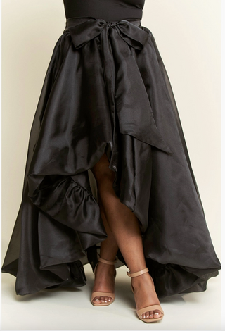 A model wearing a high-low plus sized black skirt with dramatic ruffles, featuring a tie waist and cascading layers. 