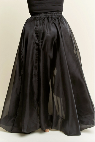 A model wearing a high-low plus sized black skirt with dramatic ruffles, featuring a tie waist and cascading layers. 