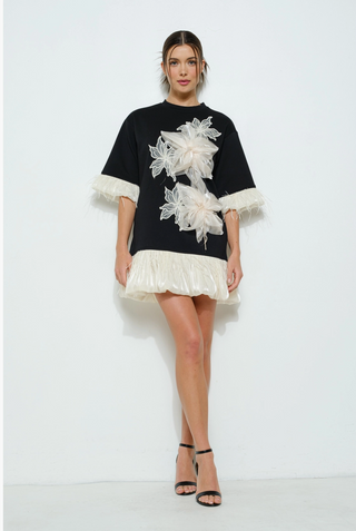 A woman wearing a black mini dress with ivory floral applique and voluminous organza bows, featuring feather-trimmed sleeves and a balloon hem.