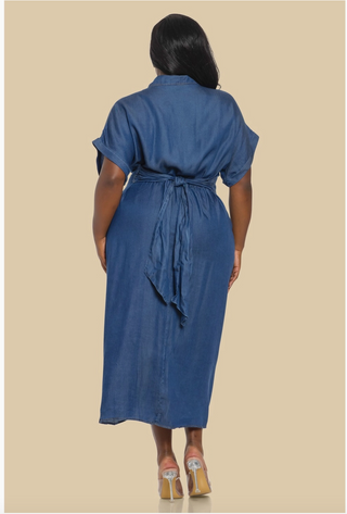 A woman confidently wearing the Denim Diva Dress, a mid-length button-down denim dress with ruched detailing at the waist, short rolled sleeves, and a front slit, paired with clear heels. The dress accentuates her curves, creating a chic and stylish look.