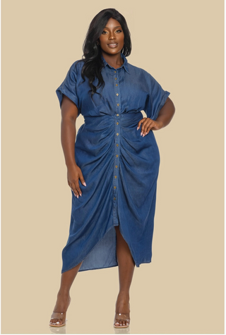 A woman confidently wearing the Denim Diva Dress, a mid-length button-down denim dress with ruched detailing at the waist, short rolled sleeves, and a front slit, paired with clear heels. The dress accentuates her curves, creating a chic and stylish look.