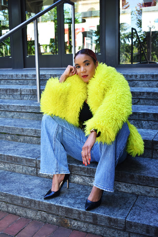 Woman, seated on steps, wearing a bright yellow faux fur jacket and straight leg blue jeans.