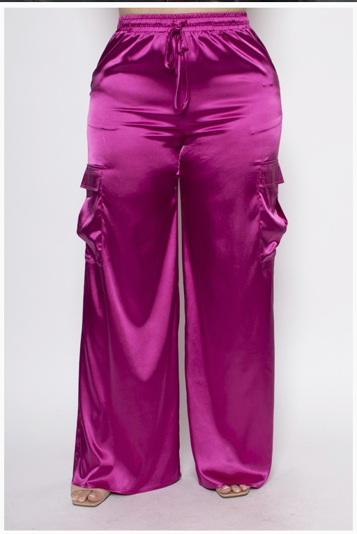 A woman wearing a pair of vibrant pink, high-waisted satin plus sized cargo pants with functional pockets and an adjustable drawstring waist.