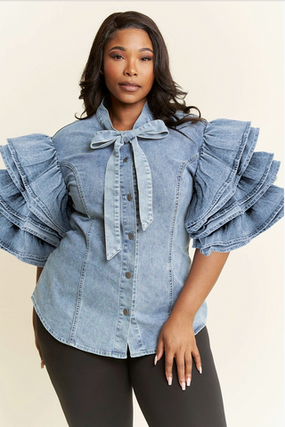 A woman wearing a plus sized blue denim shirt with tiered ruffle sleeves and a bow detail at the neckline, paired with black fitted pants.
