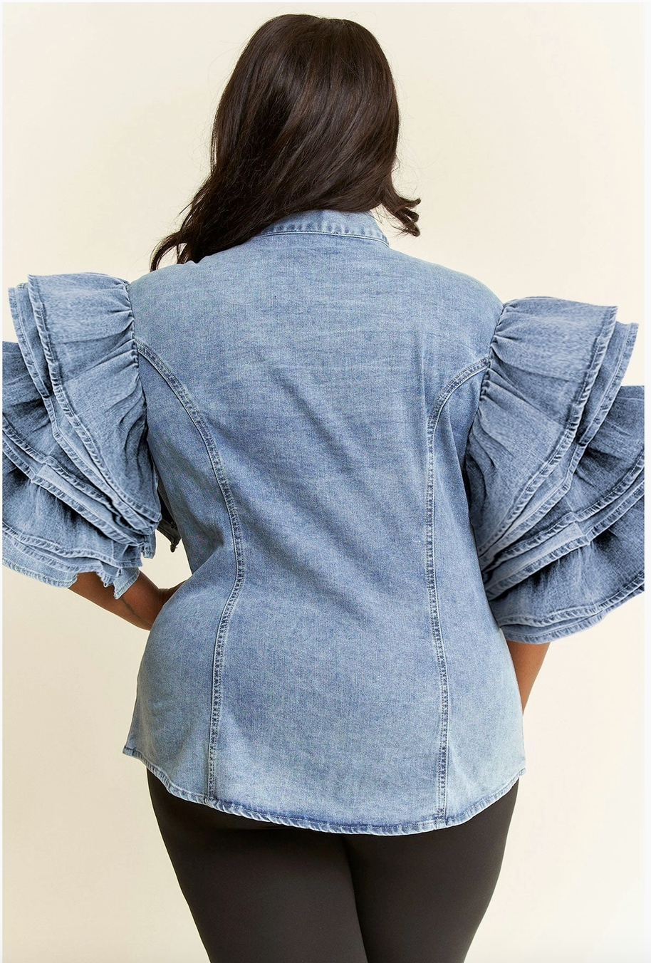 The back of a woman wearing a plus sized blue denim shirt with tiered ruffle sleeves and a bow detail at the neckline, paired with black fitted pants.
