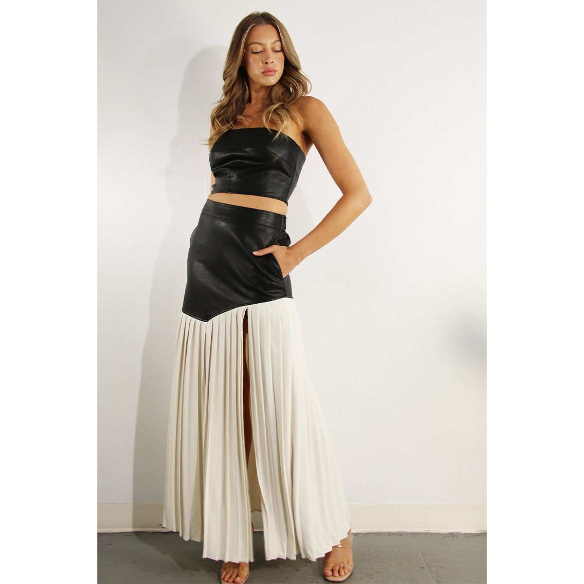 Woman wearing a two piece skirt set. The top is a faux leather tube top, and the skirt is faux leather with cream pleats. 