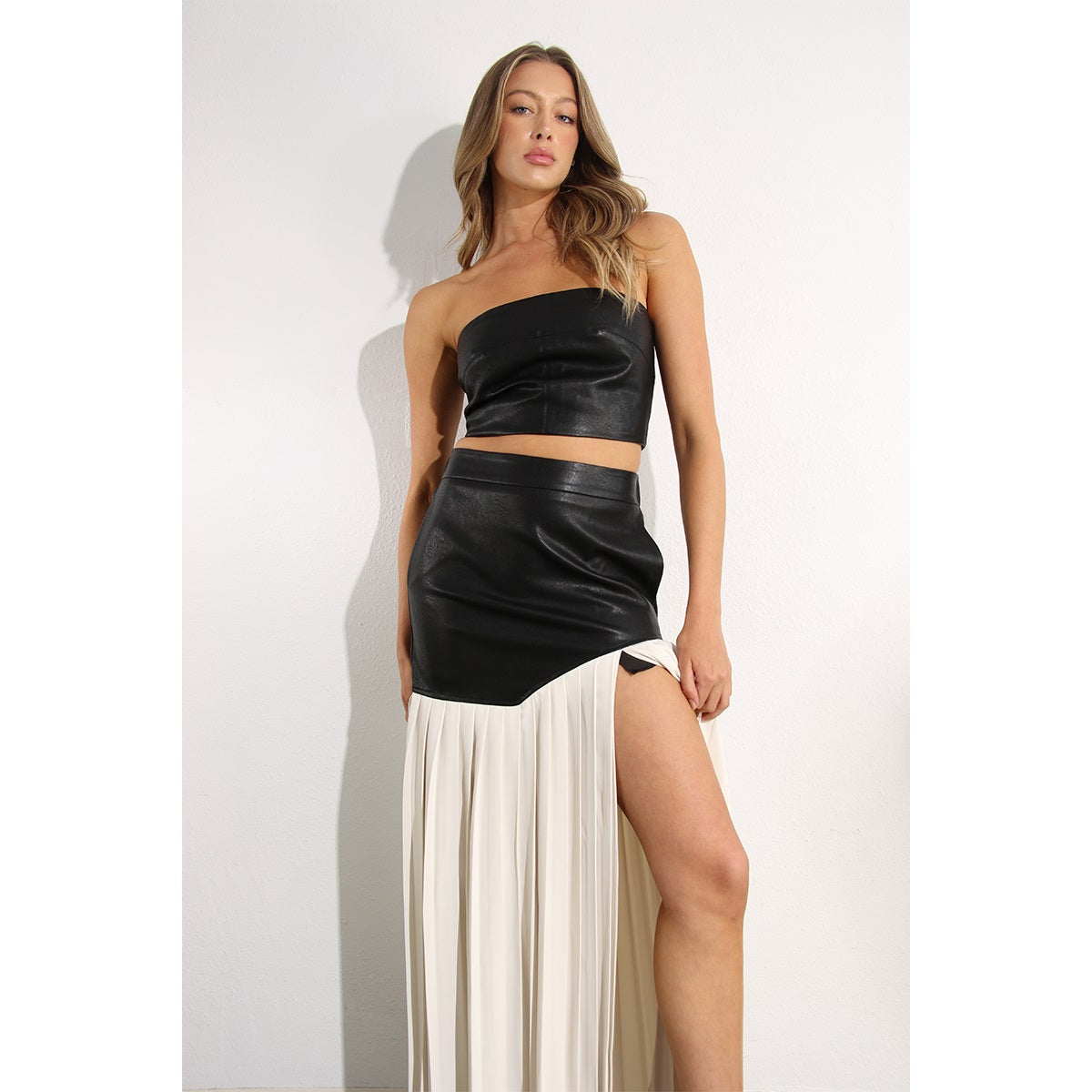 Woman wearing a two piece skirt set. The top is a faux leather tube top, and the skirt is faux leather with cream pleats. Her leg is stretched out to show off the split in the skirt.