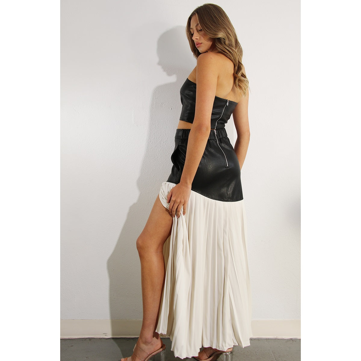 The back of a woman wearing a two piece skirt set. The top is a faux leather tube top, and the skirt is faux leather with cream pleats. 