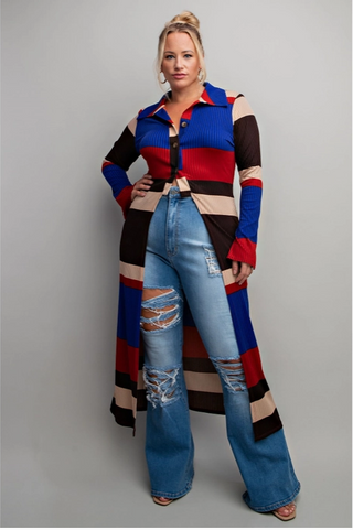 Woman wearing a longline, colorblock maxi shirt with bold horizontal stripes in blue, red, beige, and black, paired with distressed high-waisted jeans and heels. The shirt features a button-up front and is styled as a duster, creating a chic and powerful look.