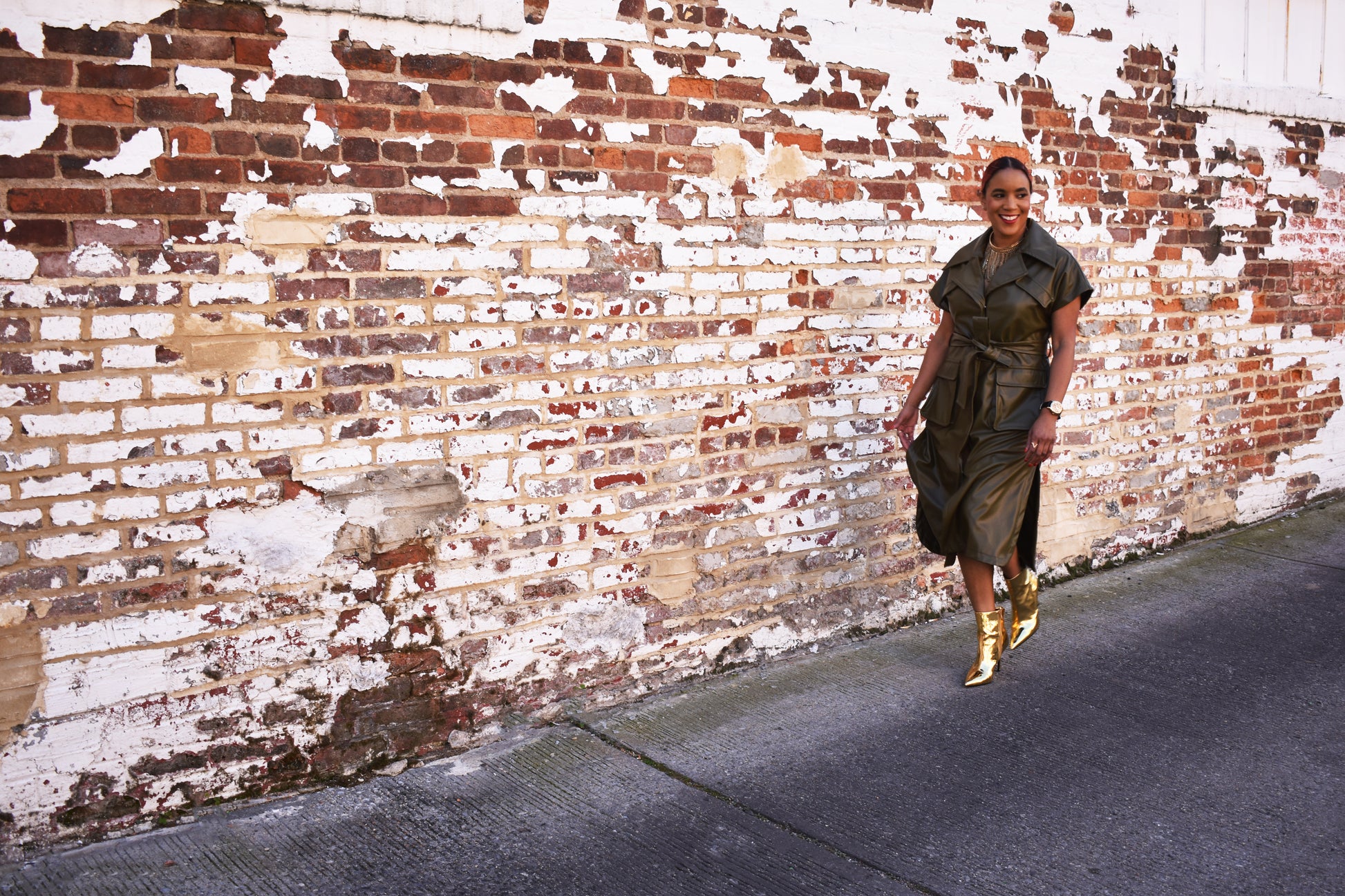 A woman wearing a green vegan leather jacket with short sleeves and oversized lapels as a dress. Paired with gold booties.