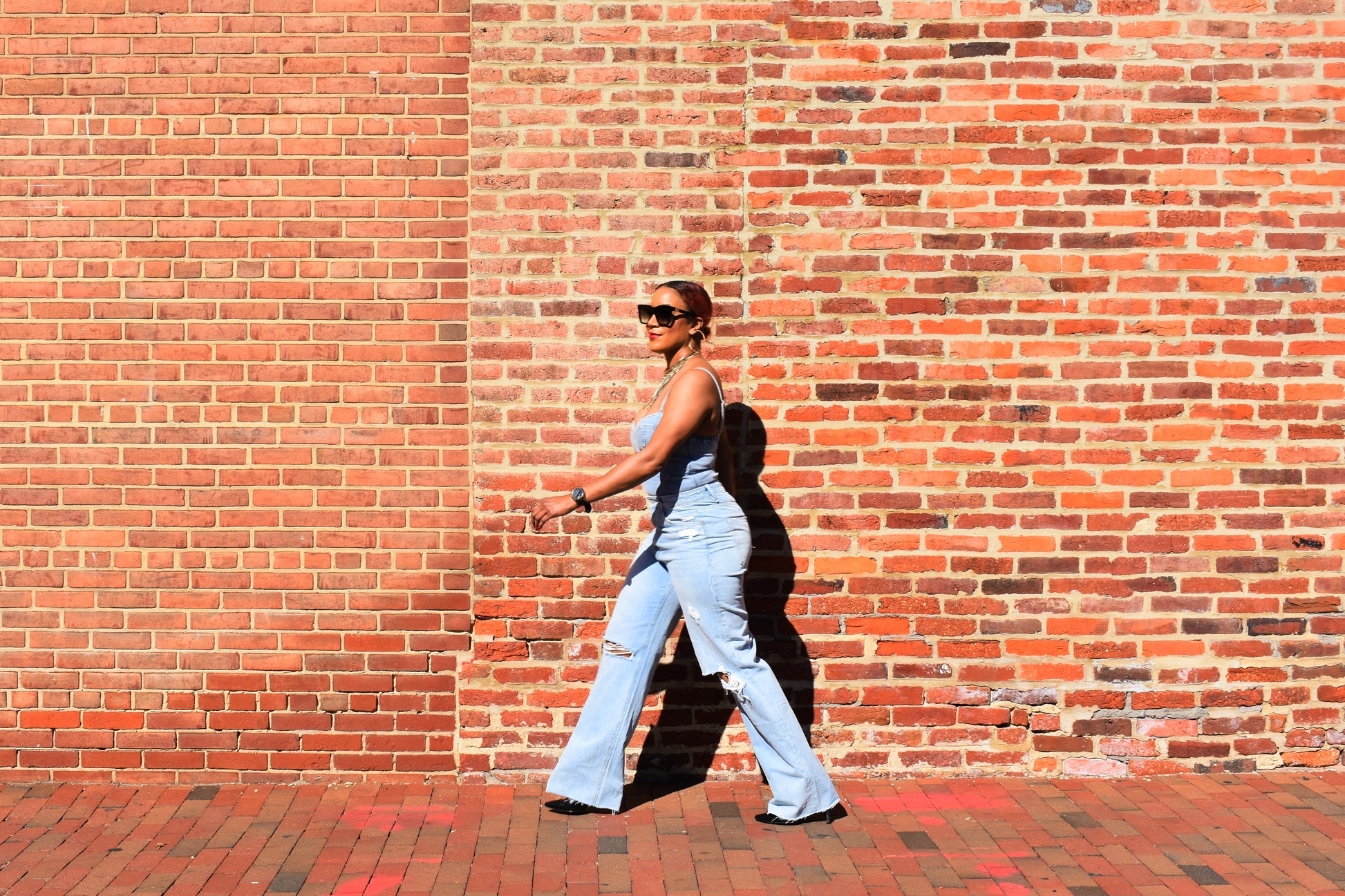 Woman wearing Split Knee Straight Leg Jeans with a denim spaghetti strap bodysuit and a stylish, chunky necklace walking against a brick wall.